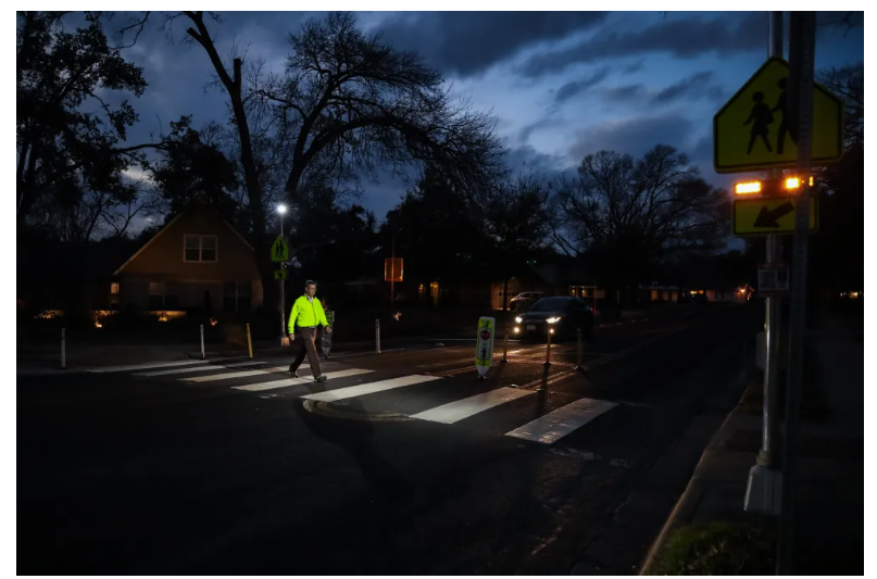 A pedestrian crossing the street, being illuminated by flood lighting attached to Rectangular Rapid Flashing Beacon.